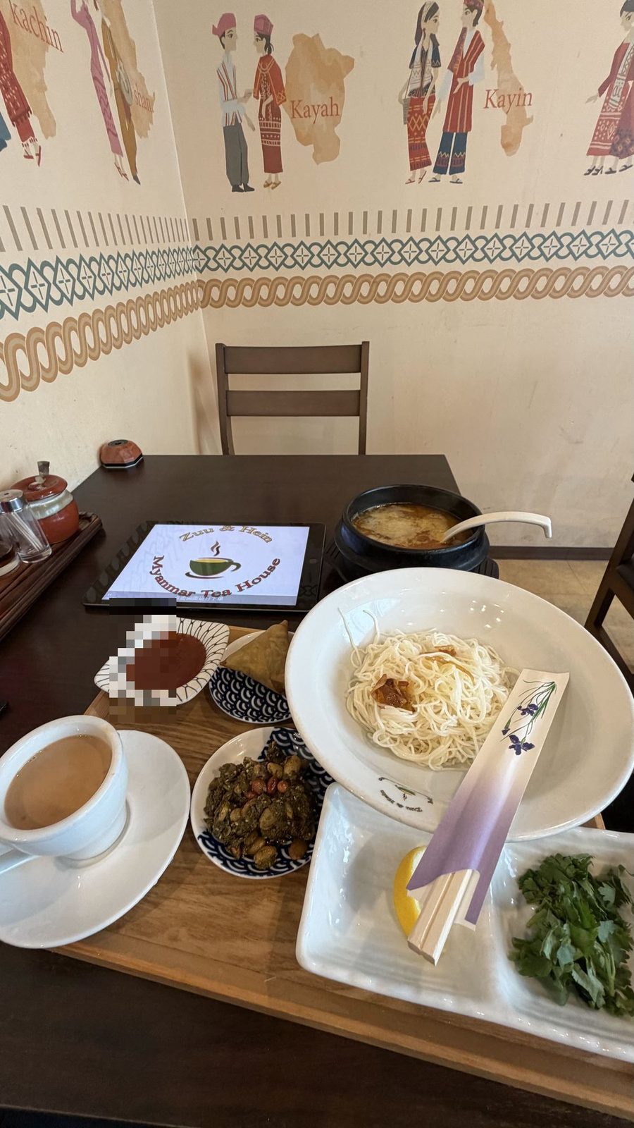 Zuu&Hein Myanmar Tea House のモヒンガーセット。石鍋のスープ、白い皿の細い麺、揚げ物や和え物の小皿、ミルクティー色の飲み物